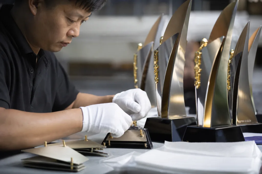 Metal Trophy Design Choices That Drive Cost, Weight, and Manufacturability 3 Worker assembling multi-part metal awards with packaging materials on a factory bench